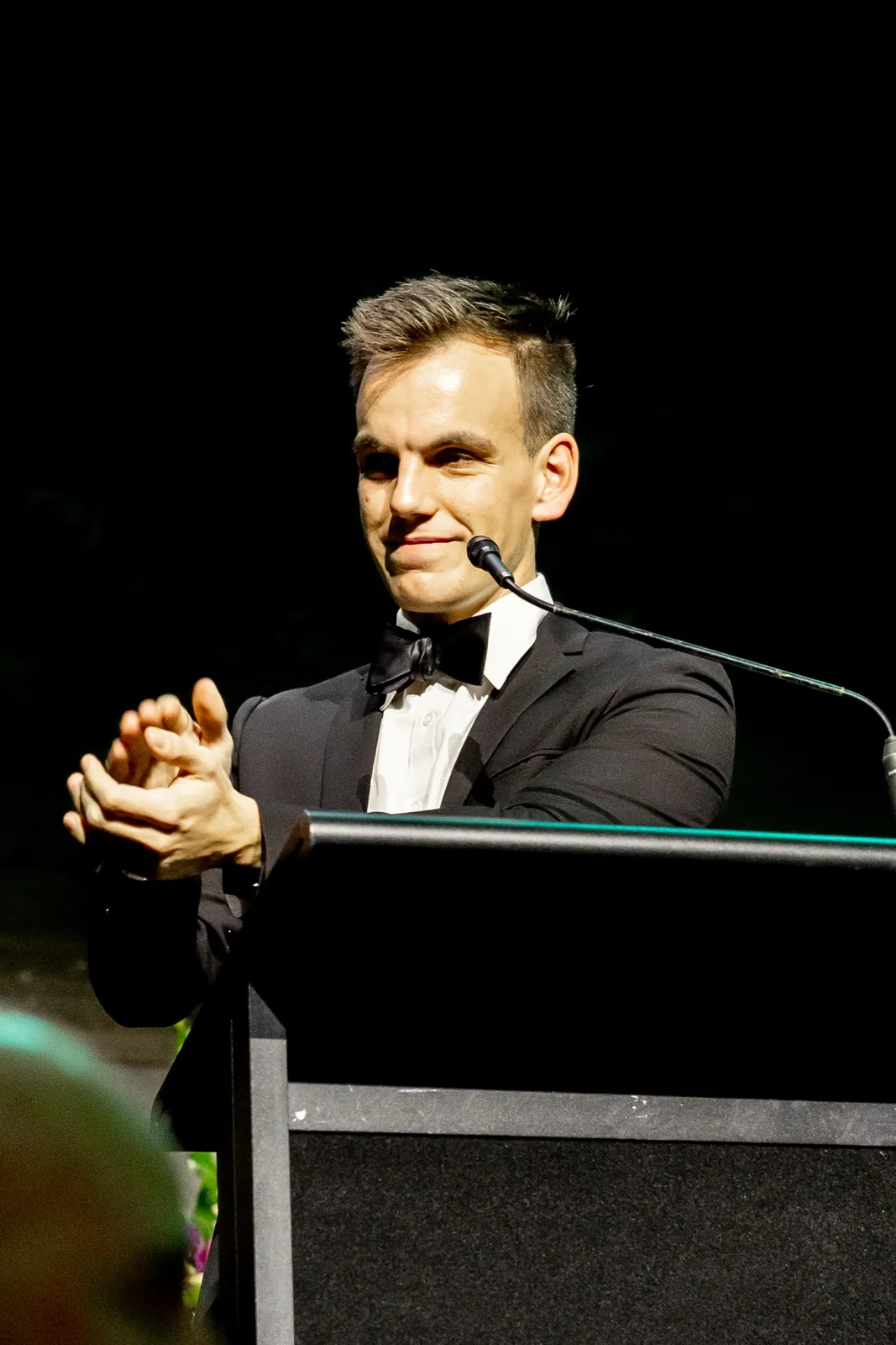 Alex applauding from the podium at the 2026 Feast To Feed gala